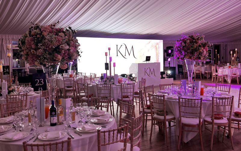 A wedding reception with pink floral décor on round tables and a lit up DJ booth.