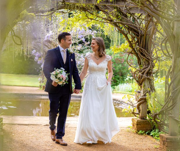 A bride and groom walking beneath the wisteria in the Pleasure Gardens. 
