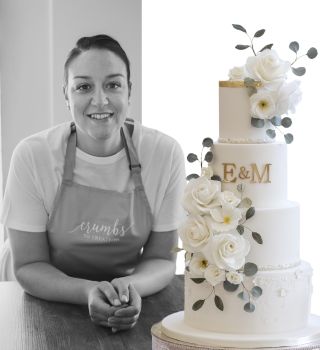 Black and white photo on a woman smiling at the camera while wearing an apron. Next to her is a cake with white roses.