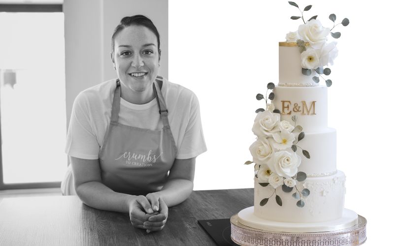 Black and white photo on a woman smiling at the camera while wearing an apron. Next to her is a cake with white roses.